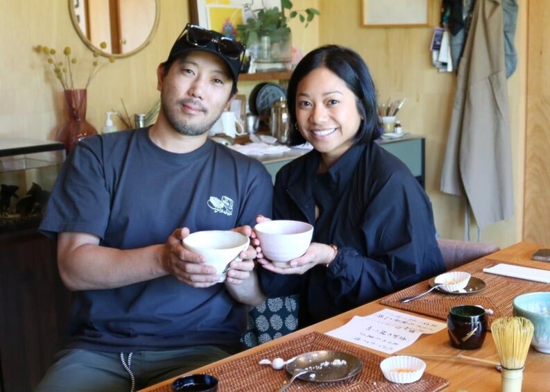 Two people holding bowls, a table with chopsticks, a whisk, and a teapot. 3