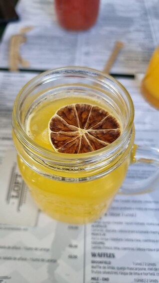 A glass jar with a dried orange slice on top, filled with a yellow liquid, placed on a menu. 1