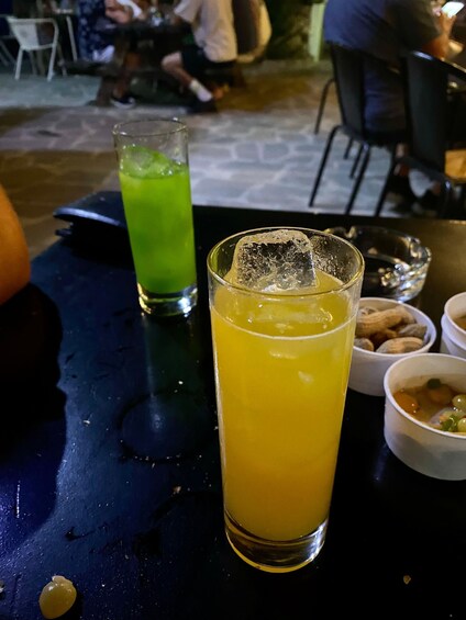 Two glasses of beverages on a table, one with a green drink and the other with a yellow drink, with ice cubes. 2