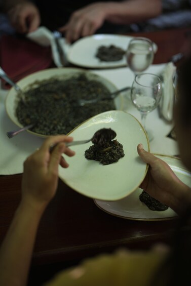 Una persona tiene un piatto con un cibo scuro, seduta a un tavolo da pranzo con altri piatti e un bicchiere. 2
