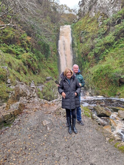 Two people standing in front of a waterfall in a forested area. 4