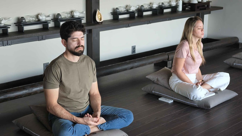 Two people sitting on meditation cushions in a room with a shelf of teapots. 2