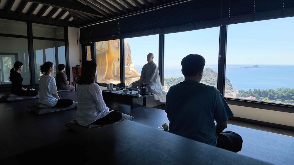 A group of people meditating in a room with large windows overlooking a coastal landscape. 4