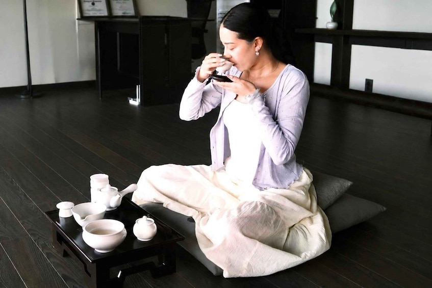 A person sitting on a cushion, holding a smartphone. There are teacups and a teapot on a tray in front of them. 3