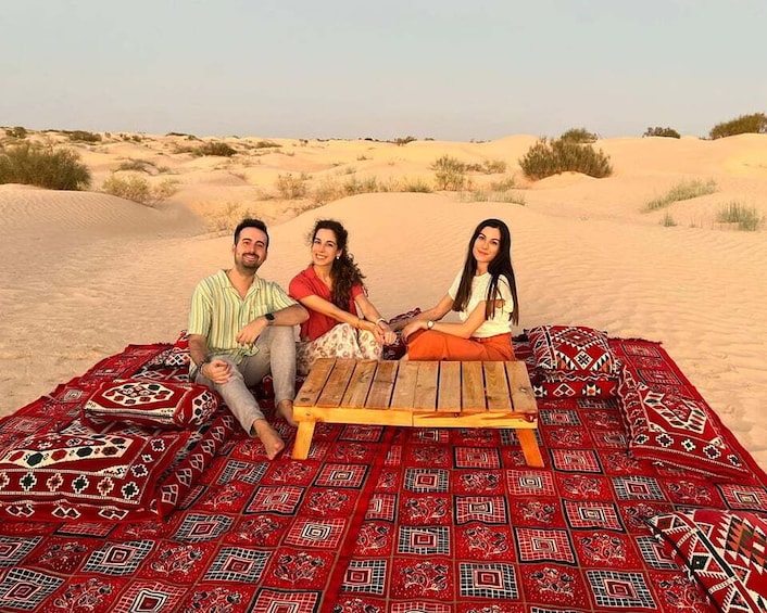 Three people sitting on a blanket with a wooden table in a desert landscape. 1