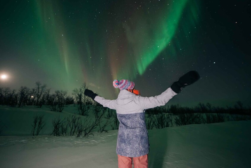 A person standing in the snow with arms raised, facing the night sky with visible aurora borealis. 4