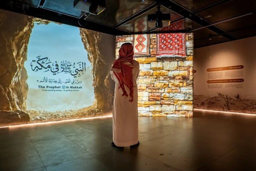 A person standing in front of a projection screen with Arabic text, a stone wall, and a red patterned cloth. 1