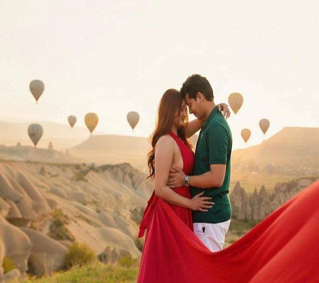 A couple embracing with a red dress flowing in the wind, set against a backdrop of a mountainous landscape and hot air balloons. 3