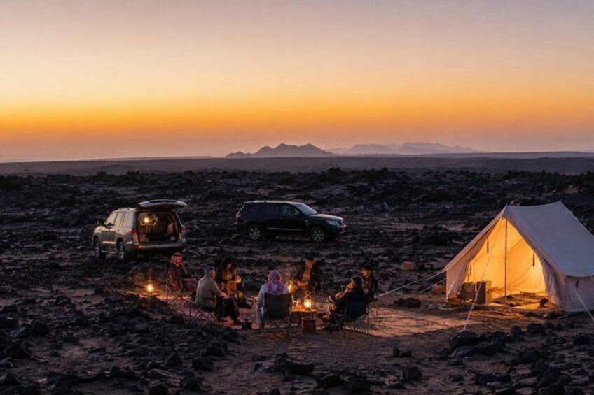 A group of people camping in a desert at sunset with vehicles and a tent. 1