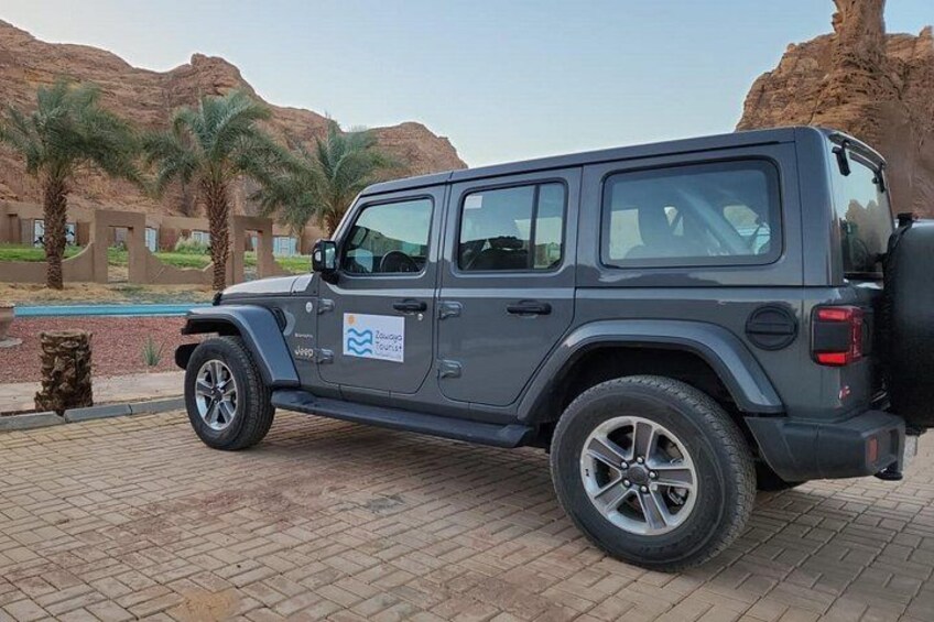 Een donkergrijze Jeep staat op een verhard gebied, met palmbomen en een rotsachtig landschap op de achtergrond. 4