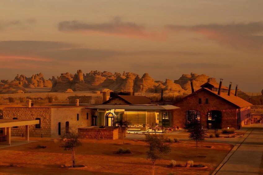A rustic building with a glass facade and a brick structure, set against a backdrop of rocky hills and a sunset sky. 1