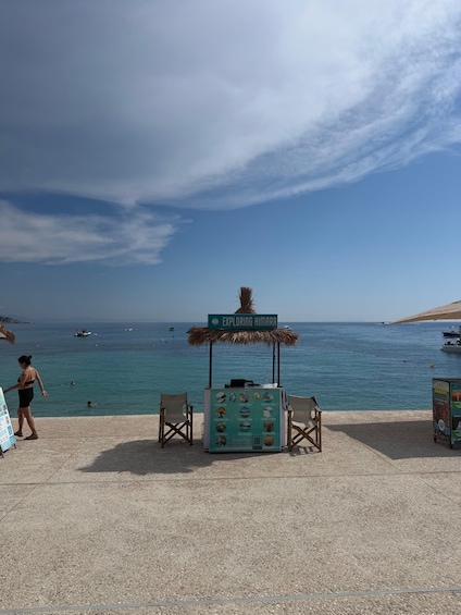 A beach with a sign reading 'Exploring Himara,' a kiosk with food options, and a clear blue sea with boats. 2