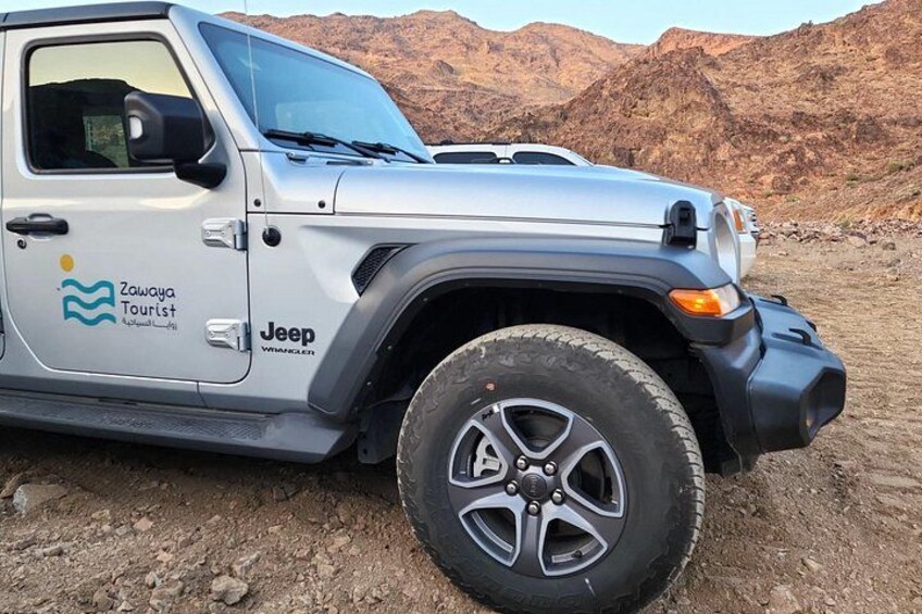 A silver Jeep Wrangler with off-road tires parked on a rocky terrain. 5