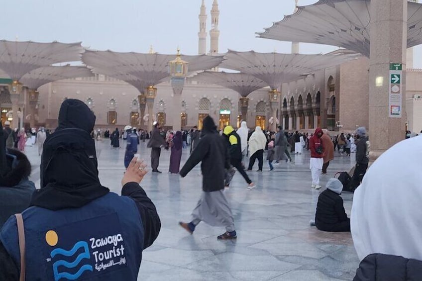 A crowded square with people walking and a large building with arches and a minaret in the background. 1