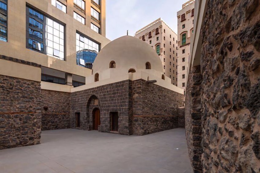 A courtyard with a central dome structure, stone walls, and modern buildings in the background. 3