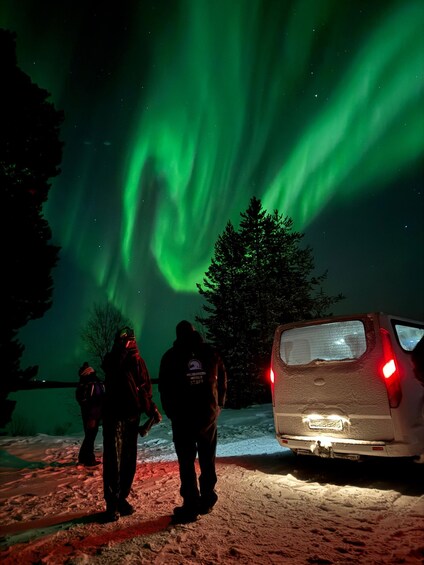People observing the Northern Lights in a snowy landscape with a car nearby. 1