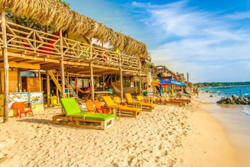 A beach with wooden structures and thatched roofs, wooden lounge chairs, and a clear blue sky. 4