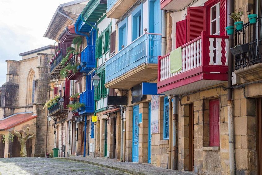 A cobblestone street lined with colorful buildings, balconies with plants, and a historic stone building in the background. 3