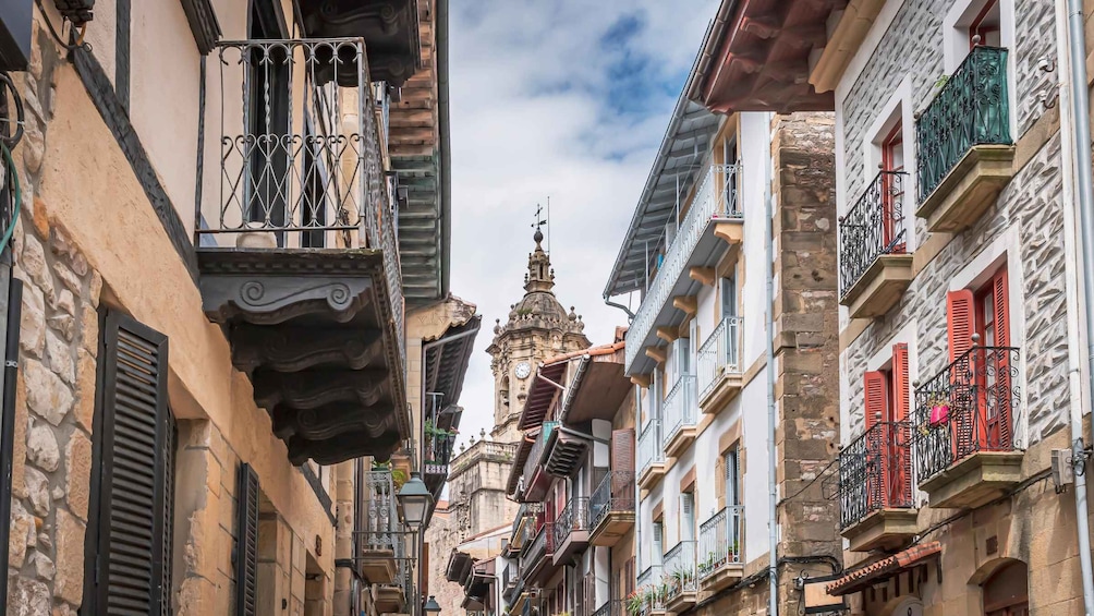 A narrow street with historic buildings, ornate balconies, and a prominent clock tower in the background. 2