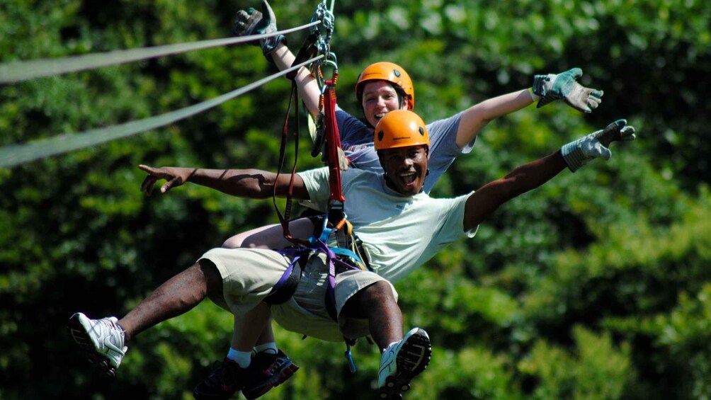 Two people ziplining, wearing helmets and harnesses, with trees in the background. 1
