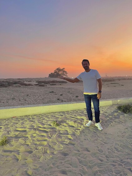 A person standing on a sandy path with footprints, a tree, and a sunset in the background. 5