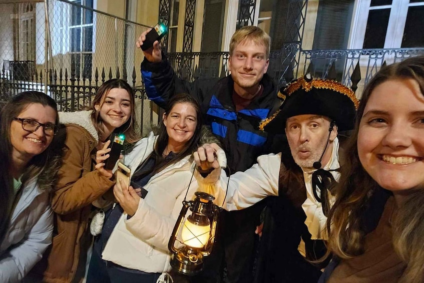 A group of people in a festive outdoor setting, some holding a lantern and a bottle, with a metal fence and buildings in the background. 1