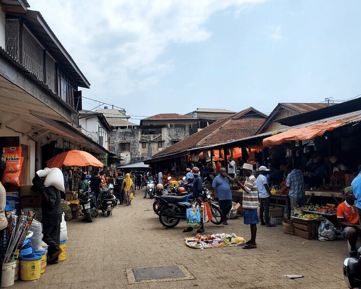 A bustling market with vendors and shoppers, motorcycles parked along the sides, and various goods displayed on tables. 2
