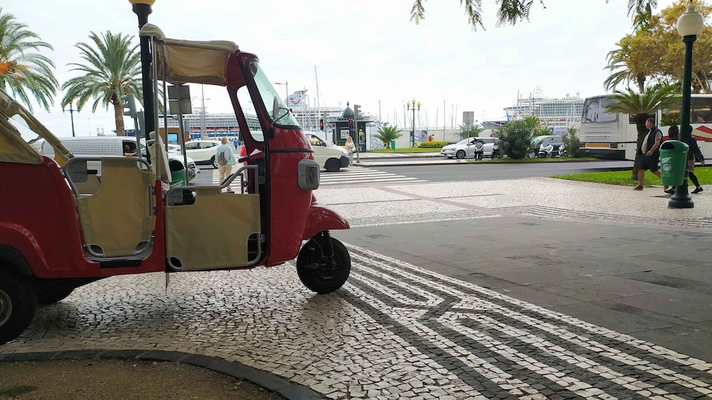 A red three-wheeled vehicle parked on a cobblestone street with palm trees and a cruise ship in the background. 2