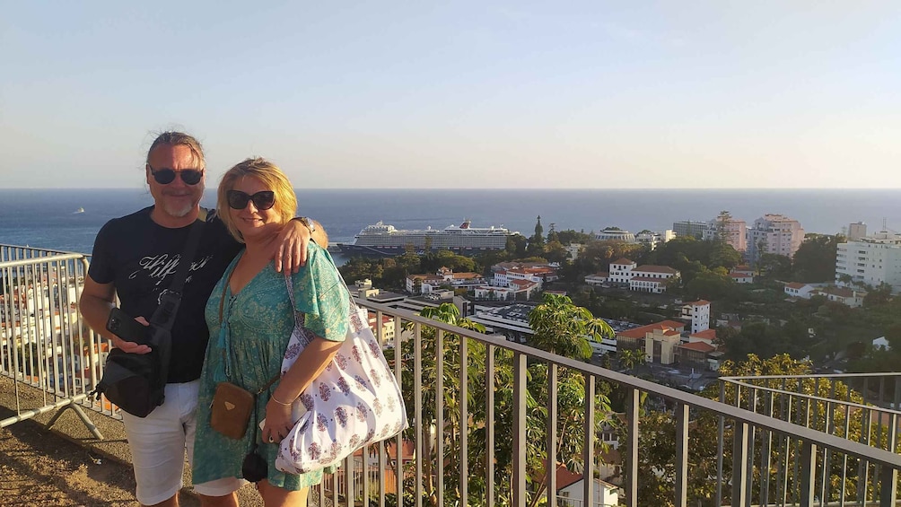 A couple standing on a balcony overlooking a coastal cityscape with buildings and a cruise ship in the distance. 4