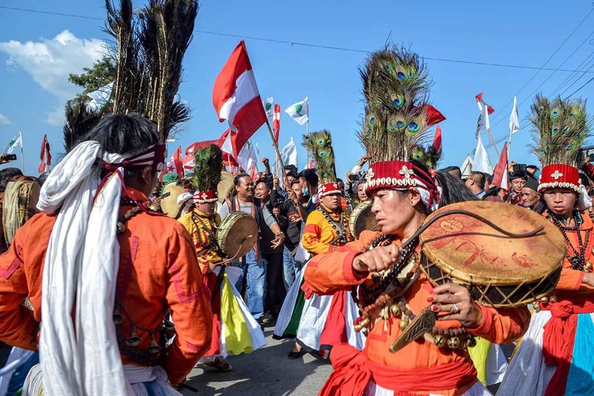 A group of people in traditional attire, some holding flags, and others playing drums, indicating a cultural or festive event. 1