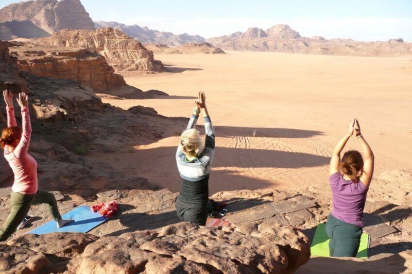 Three people practicing yoga on mats in a desert landscape with rocky terrain and mountains in the background. 2
