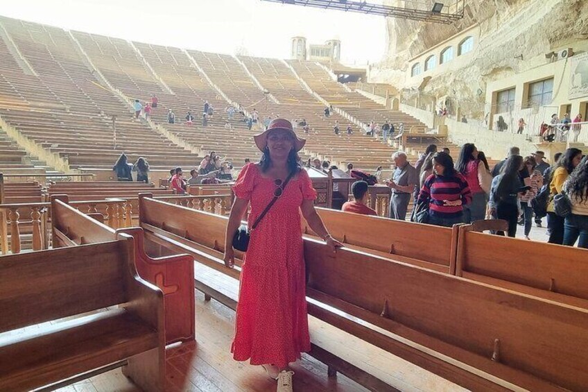 A person in a red dress standing in an auditorium with wooden pews and a stone wall. 5