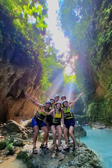 A group of people in helmets and life vests posing on a rocky outcrop by a clear water stream in a forested area. 5