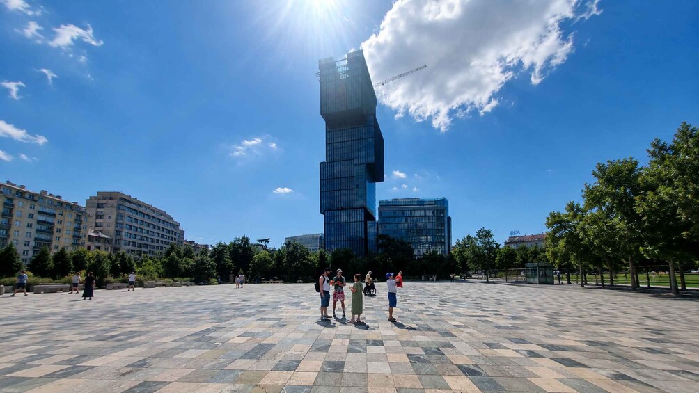 A plaza with a modern glass building, people walking, and surrounding trees. 2