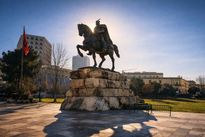A statue of a figure on horseback on a pedestal in a park with buildings in the background. 1