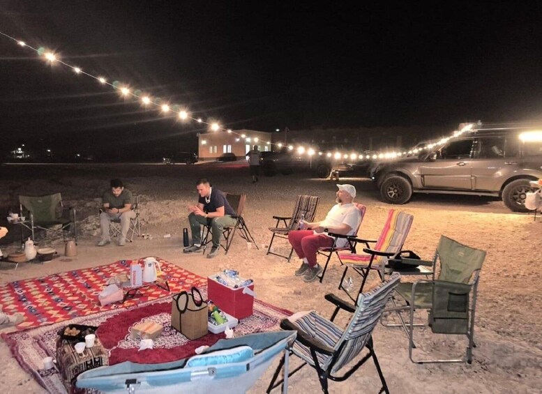 A group of people sitting on chairs and blankets on a sandy beach at night, with string lights overhead. 1