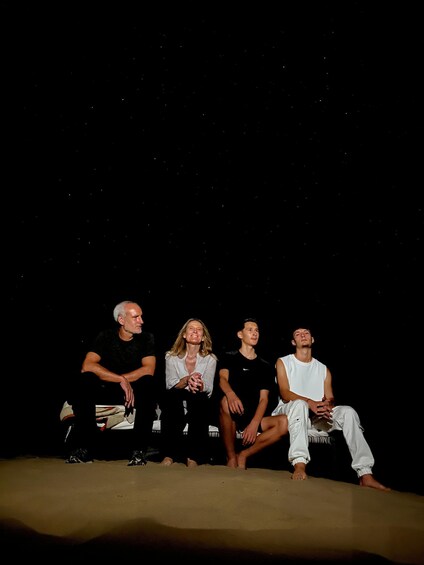 Four people sitting on a sandy surface against a dark sky with stars. 4