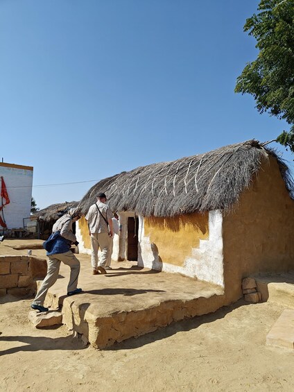 A traditional mud house with a thatched roof, a person taking a photo, and another person walking towards the entrance. 2