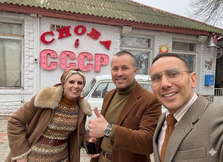 Three people in front of a building with a sign reading "CHODA CCCP". 1