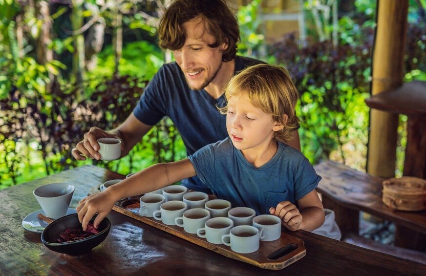 Dos personas preparando té en una mesa de madera al aire libre, con follaje en el fondo. 5