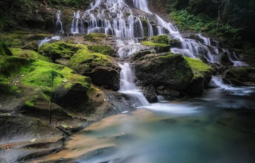 A multi-tiered waterfall cascading over moss-covered rocks in a forested area. 2
