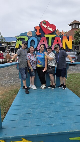 A group of people standing in front of a colorful sign with the word 'ROATAN' and a heart-shaped sign with 'Halu Kiliki' written on it. 4