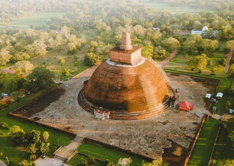 A large, ancient stupa surrounded by a square courtyard with a few scattered buildings and trees. 1