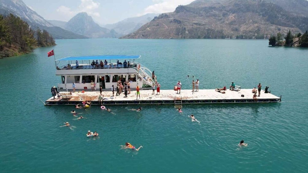 A boat docked at a pier with people swimming in the water and a mountainous landscape in the background. 4