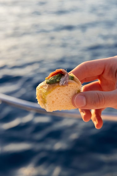 A hand holding a sandwich with a green topping, against a blurred background of water. 4