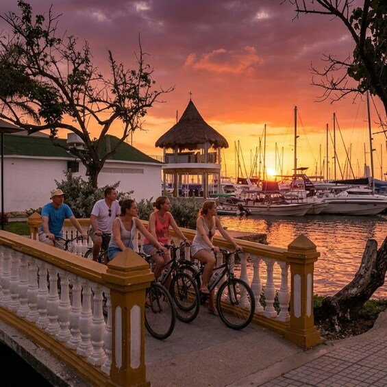 A sunset over a marina with boats, a thatched-roof gazebo, and people riding bicycles on a bridge. 3