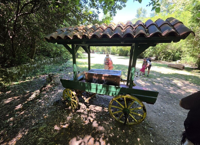 A rustic wooden cart with a tiled roof, wheels, and a bench, situated in a wooded area. 3