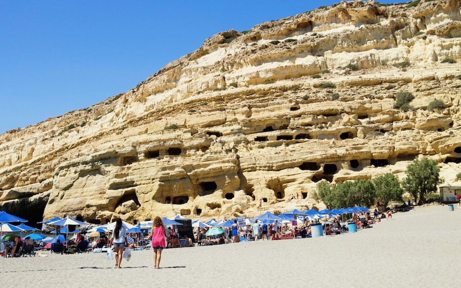 A rocky cliff with numerous caves and a beach with people and umbrellas. 4