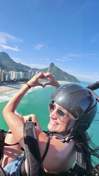 A person making a heart shape with their hands, wearing a helmet and a harness, with a coastal landscape and buildings in the background. 5