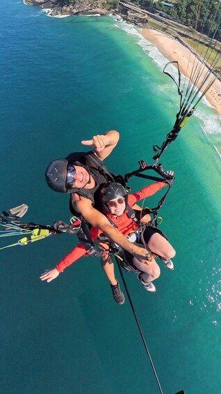 A person parasailing over turquoise waters with a beach and rocky coastline in the background. 1